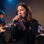 Une chanteuse répète avec un microphone à la main dans une petite salle de concert, accompagnée d’un groupe en arrière-plan sous une lumière de scène sobre.
